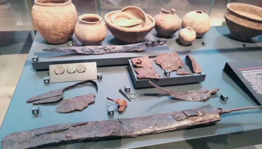 Reperti archeologici, tra cui vasi, monete e armi, simili a quelli della patera di Parabiago, esposti in un museo.