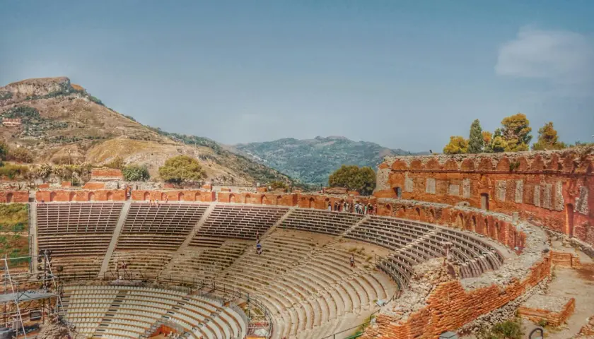 Antico teatro greco in Italia, con gradinate in pietra e rovine in mattoni rossi, incastonato tra colline verdi e un cielo azzurro.