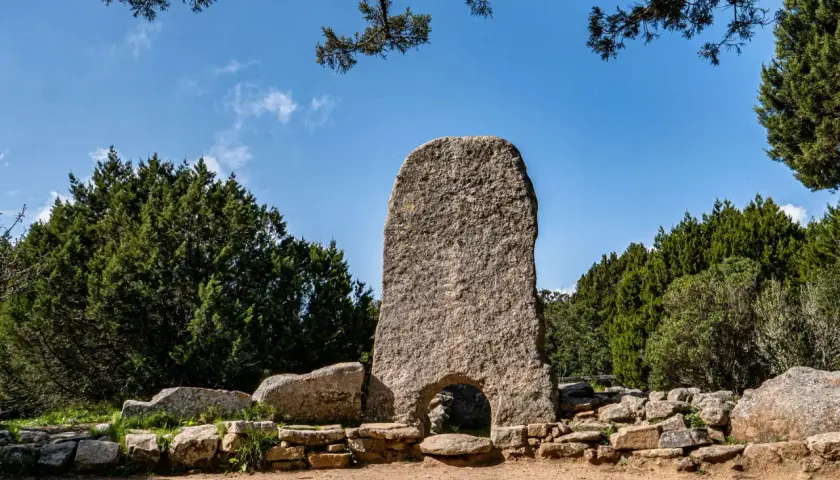 Un imponente menhir con un arco alla base, circondato da pietre antiche e vegetazione rigogliosa. Tra le tombe dei giganti più belle, questo sito archeologico incanta.