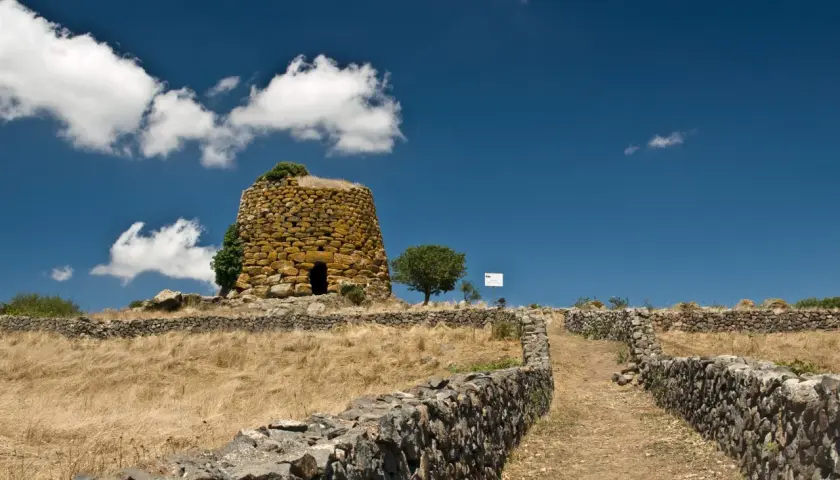 Antichi nuraghi sardi, testimoni di un passato di guerra e civiltà. Un sentiero di pietra conduce verso la torre, immersa in un paesaggio arido sotto un cielo azzurro.