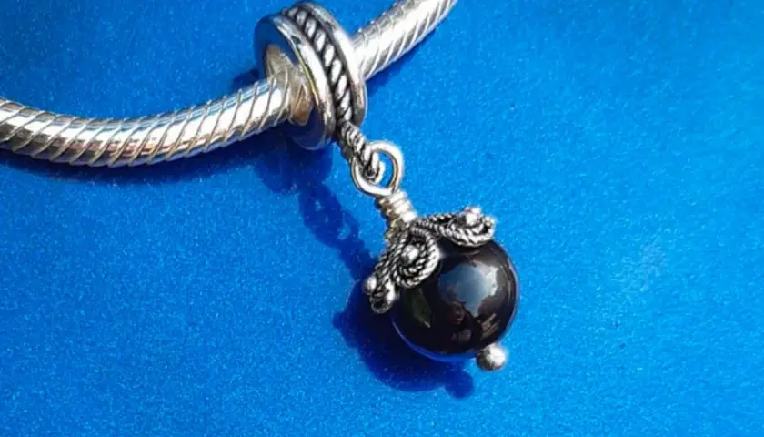 Un ciondolo con una pietra nera lucida, ispirato agli antichi amuleti sardi, pende da un bracciale d'argento su sfondo blu.