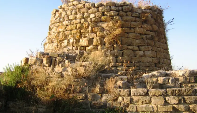 Il nuraghe Piscu, con le sue pietre antiche e l'erba secca, si staglia contro il cielo azzurro, testimone silenzioso di storie millenarie.