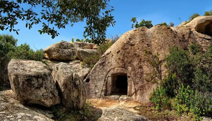 Ingresso di una delle **domus de janas**, tombe ipogeiche scavate nella roccia in Sardegna, circondata da rocce e vegetazione mediterranea.
