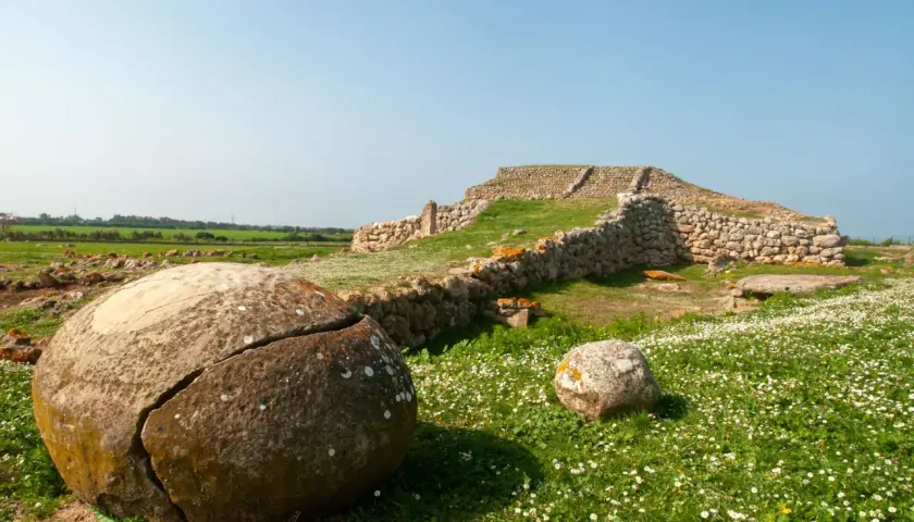 Antiche pietre e rovine di un ziqqurat in Sardegna, circondate da un prato fiorito e rocce tondeggianti.