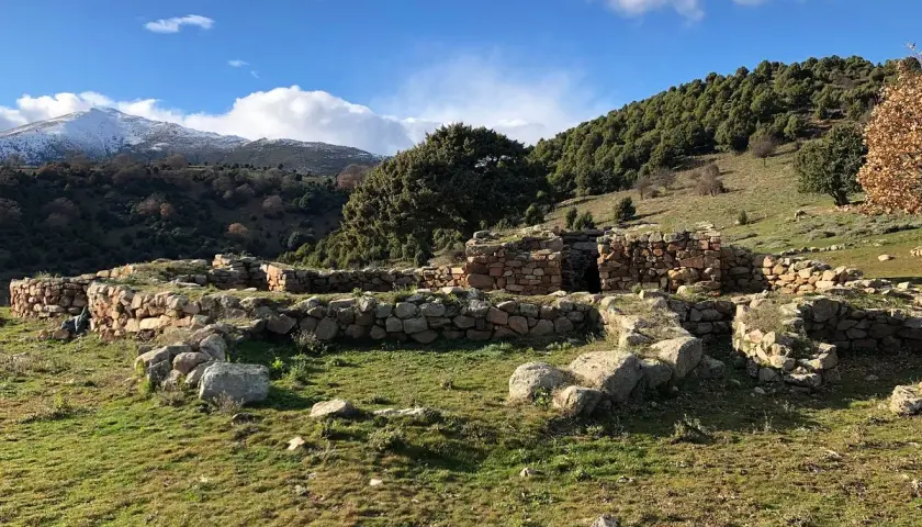 Antiche rovine di pietra, s'arcu e is forros, su un prato verde sotto un cielo azzurro con nuvole bianche e montagne innevate sullo sfondo.
