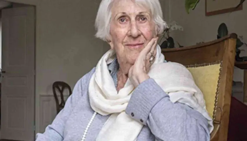 Annick de Souzenelle, con un sorriso sereno, appoggia il viso sulla mano, avvolta in una sciarpa chiara.