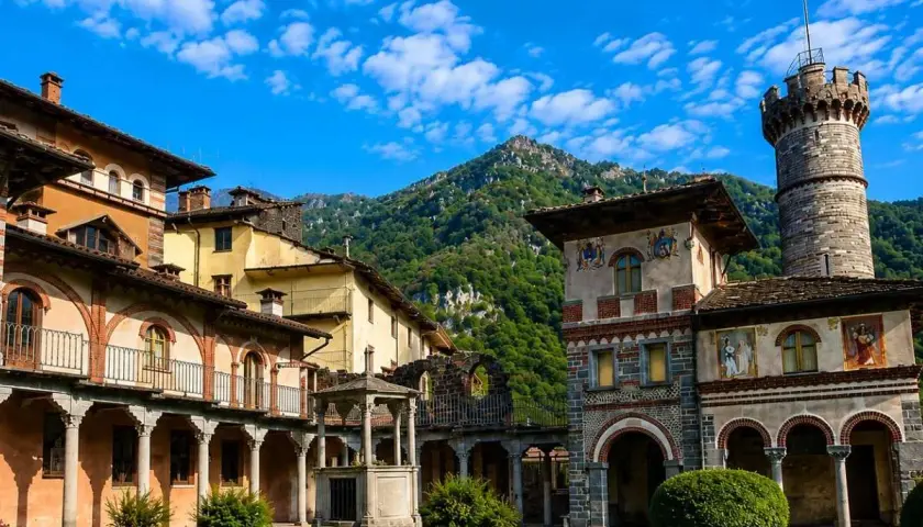 Cortile di un complesso architettonico con torre, affreschi e un pozzo, immerso nel verde. Sembra un luogo avvolto da un'aura di mistero, quasi una rosazza esoterica.