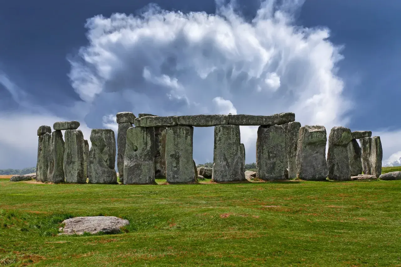 Antico cromlech di pietre imponenti su un prato verde, sotto un cielo drammatico con nuvole bianche e blu.