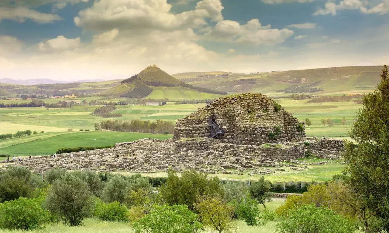 Antico nuraghe in pietra, con scale esterne, immerso in un paesaggio verdeggiante con colline ondulate e un cielo nuvoloso.