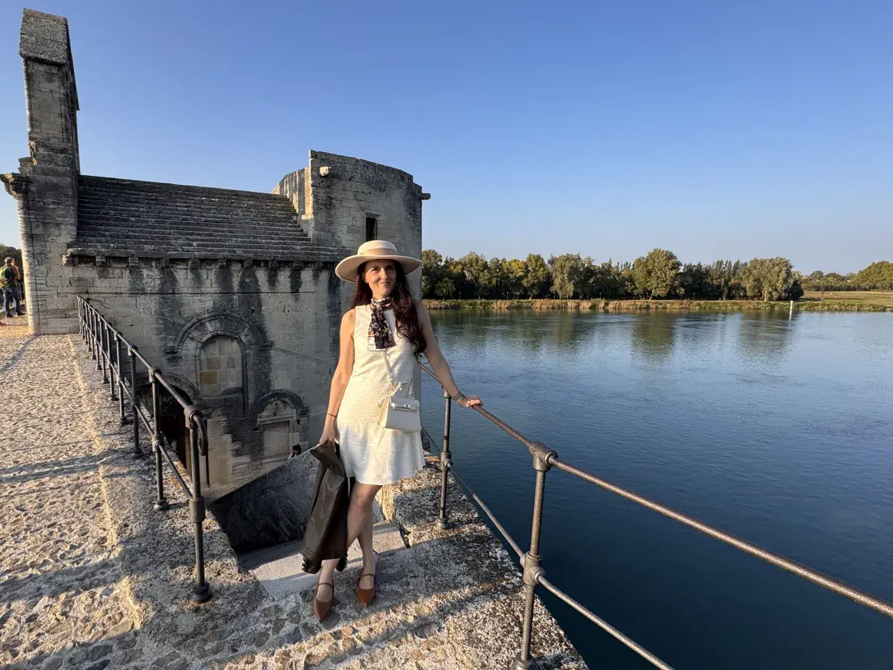 Donna con cappello e giacca in pelle, in piedi sul ponte di pietra vicino al fiume, con un edificio antico sullo sfondo, che ricorda un altare rupestre di Santo Stefano.