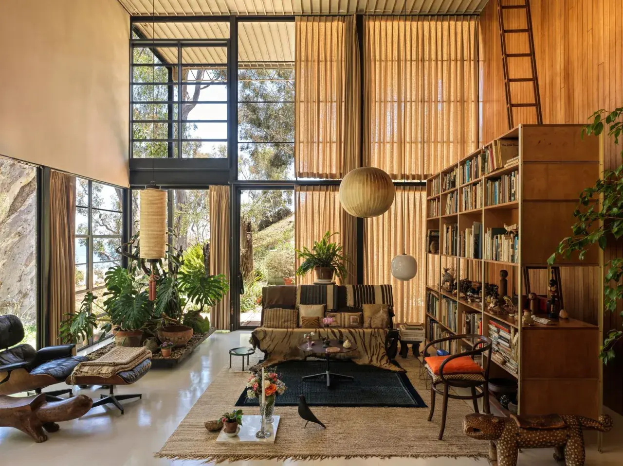 Interno della Eames House con grandi finestre, mobili iconici, piante rigogliose e una libreria piena.