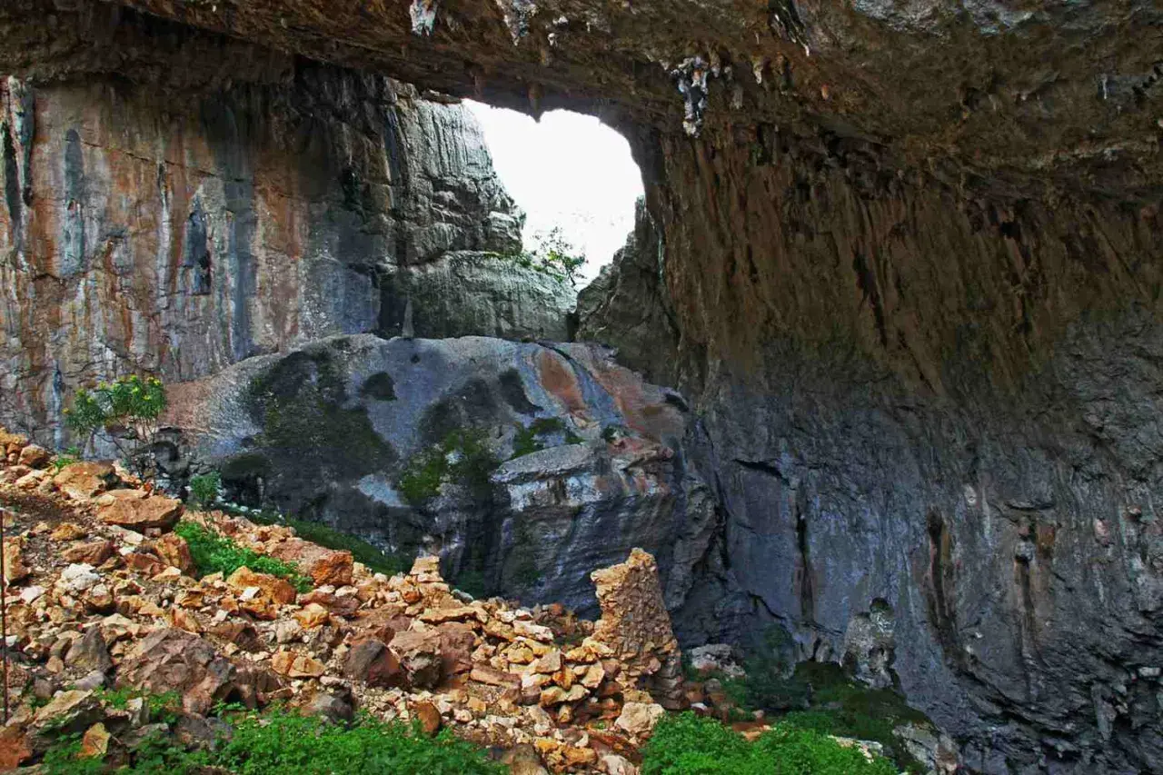 Ingresso di una grotta con rocce stratificate e vegetazione. Un arco naturale si apre verso l'esterno, rivelando un paesaggio luminoso. Un sito archeologico suggestivo.