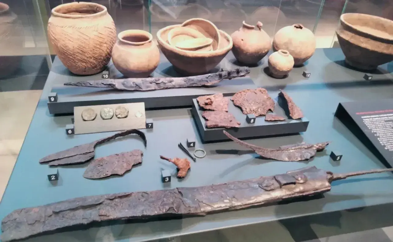 Reperti archeologici, tra cui vasi, monete e armi, simili a quelli della patera di Parabiago, esposti in un museo.