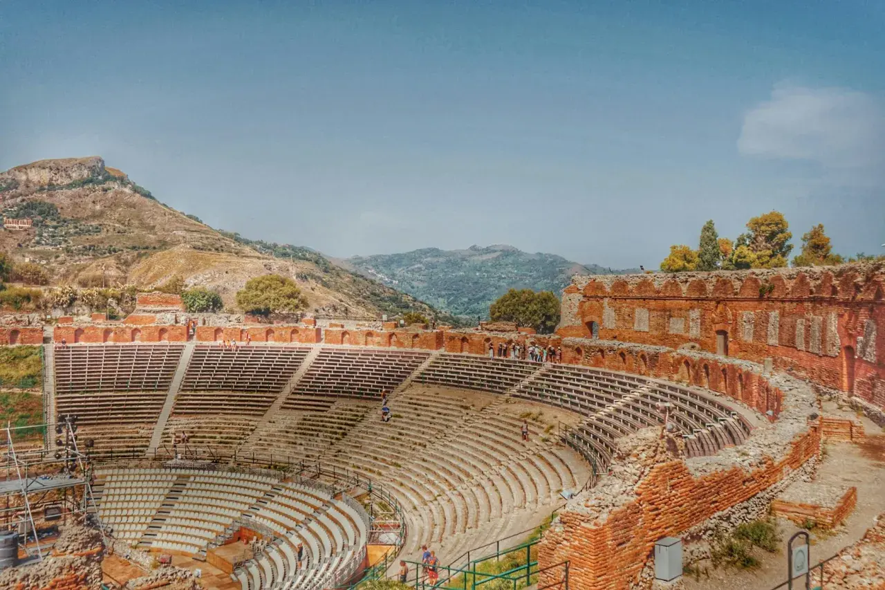 Antico teatro greco in Italia, con gradinate in pietra e rovine in mattoni rossi, incastonato tra colline verdi e un cielo azzurro.