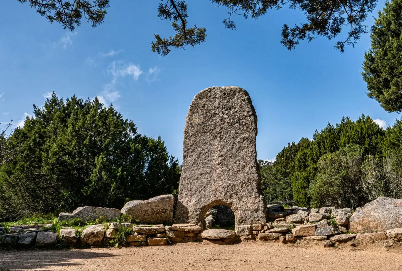 Un imponente menhir con un arco alla base, circondato da pietre antiche e vegetazione rigogliosa. Tra le tombe dei giganti più belle, questo sito archeologico incanta.