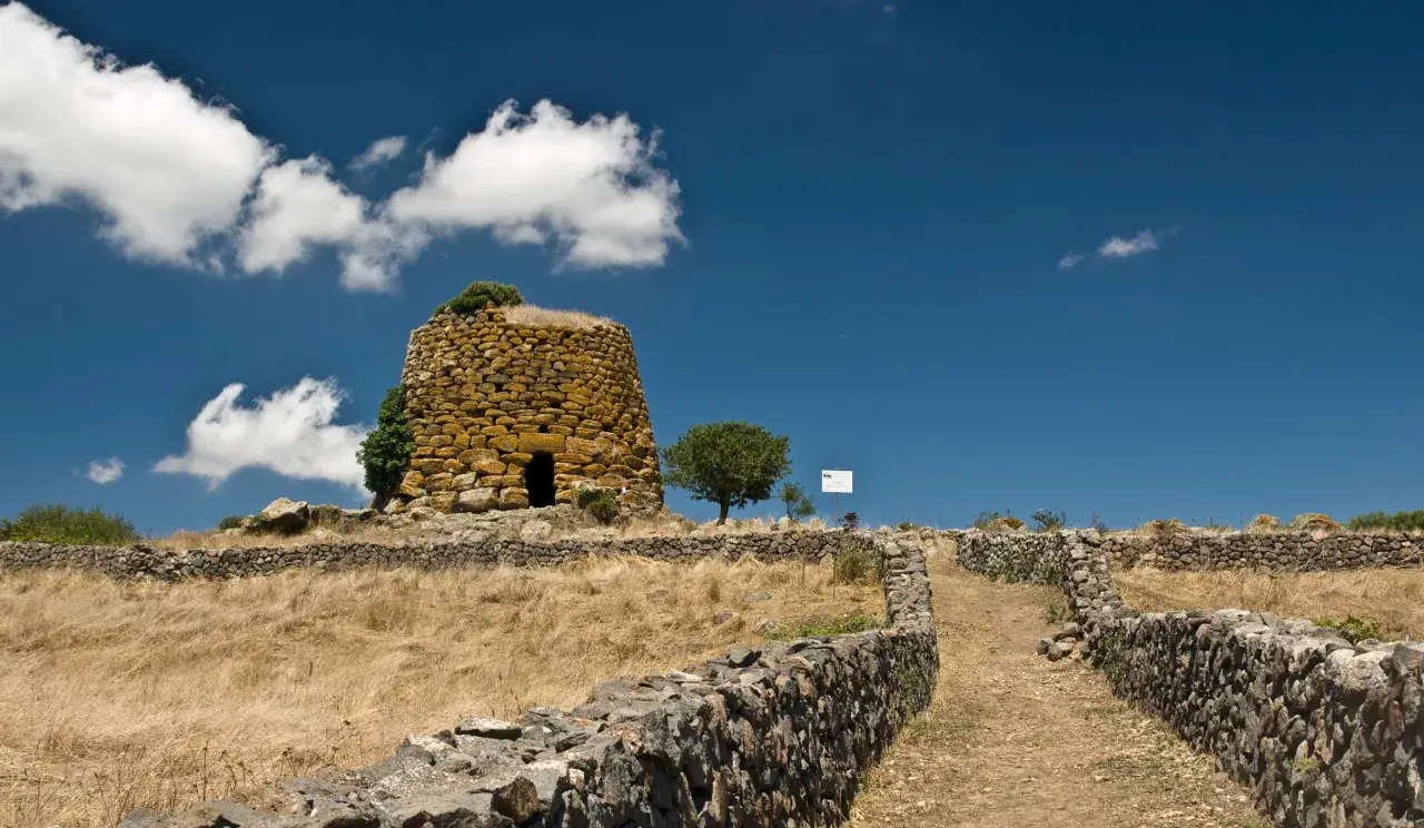 Antichi nuraghi sardi, testimoni di un passato di guerra e civiltà. Un sentiero di pietra conduce verso la torre, immersa in un paesaggio arido sotto un cielo azzurro.