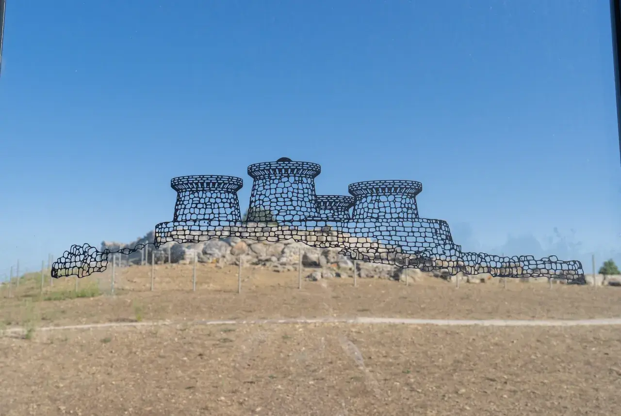 Disegno stilizzato del nuraghe Genna Maria, con torri tronche e mura merlate, su uno sfondo di cielo azzurro e terreno brullo.