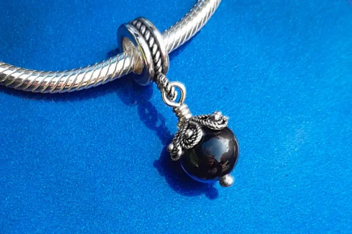 Un ciondolo con una pietra nera lucida, ispirato agli antichi amuleti sardi, pende da un bracciale d'argento su sfondo blu.