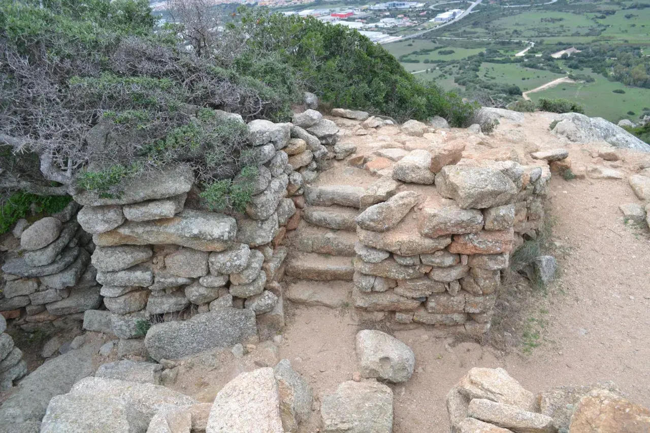 Antiche pietre del nuraghe Cabu Abbas, con gradini in pietra e vegetazione mediterranea.