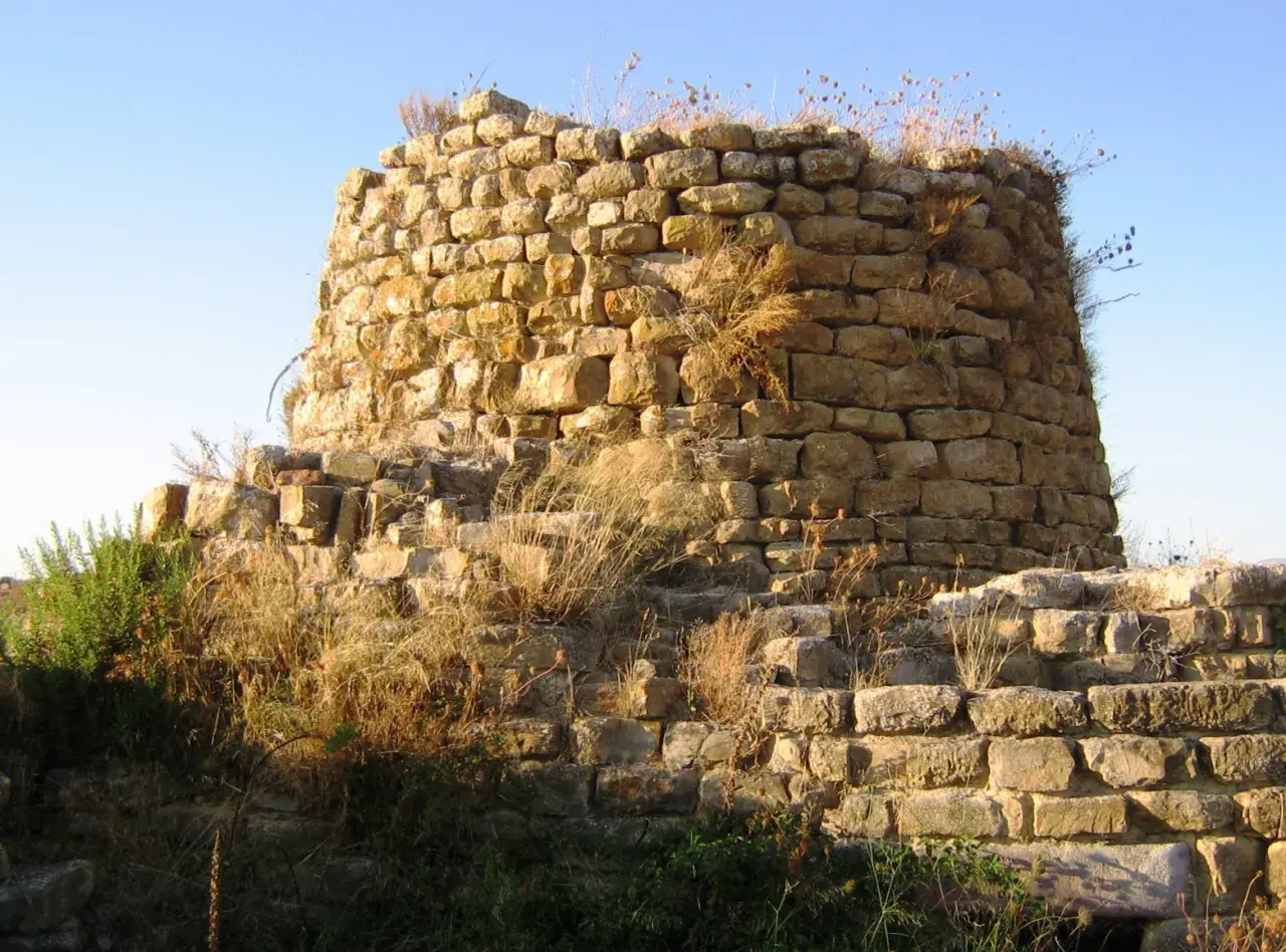 Il nuraghe Piscu, con le sue pietre antiche e l'erba secca, si staglia contro il cielo azzurro, testimone silenzioso di storie millenarie.