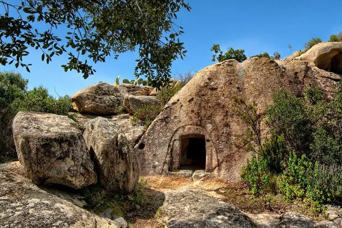 Ingresso di una delle **domus de janas**, tombe ipogeiche scavate nella roccia in Sardegna, circondata da rocce e vegetazione mediterranea.
