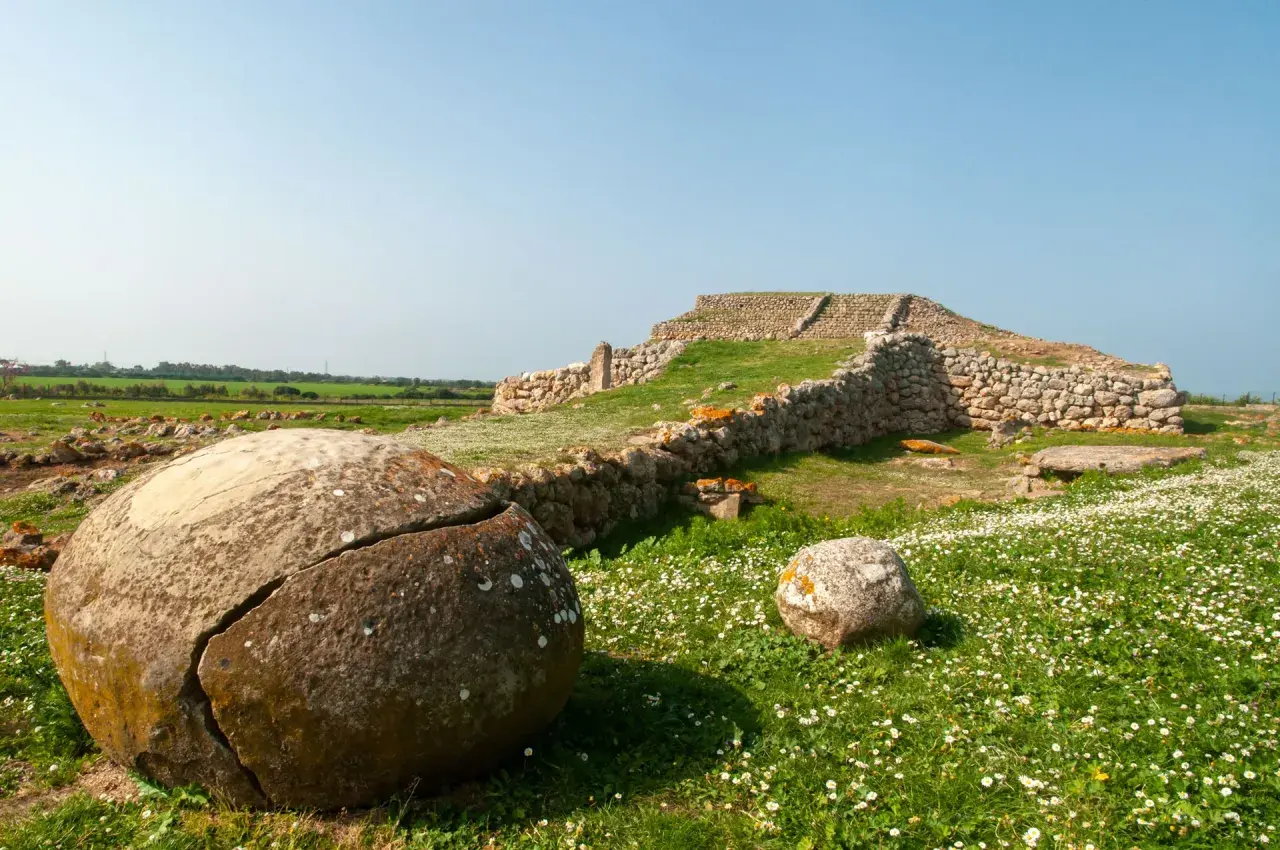 Antiche pietre e rovine di un ziqqurat in Sardegna, circondate da un prato fiorito e rocce tondeggianti.