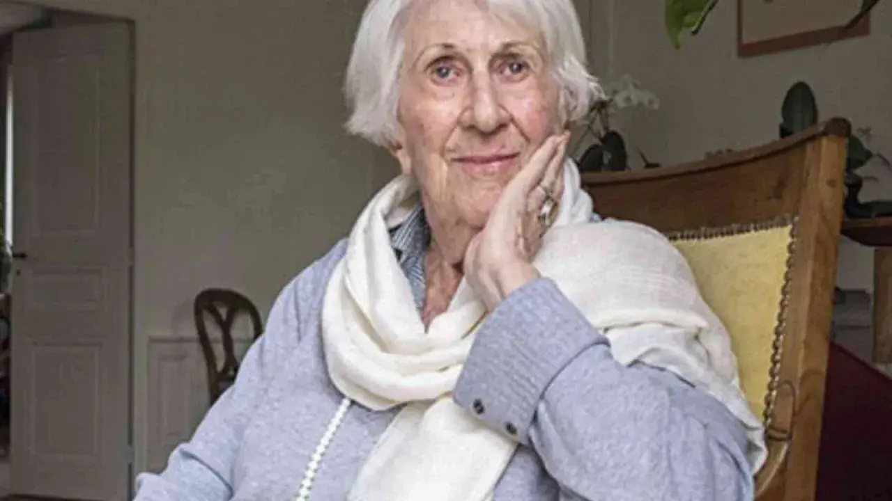 Annick de Souzenelle, con un sorriso sereno, appoggia il viso sulla mano, avvolta in una sciarpa chiara.