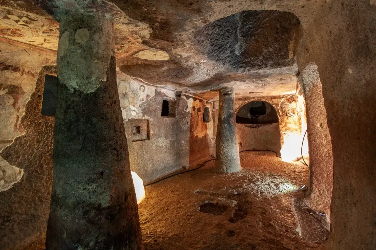 Interno di una tomba etrusca con affreschi antichi e una colonna scolpita a forma di sa pala larga.