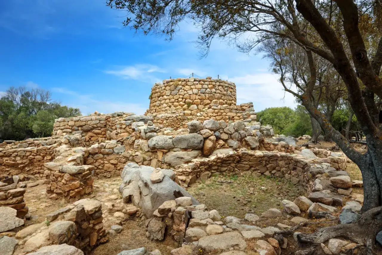Antico villaggio nuragico in pietra, con una torre centrale ben conservata e resti di mura circostanti, sotto un cielo azzurro.