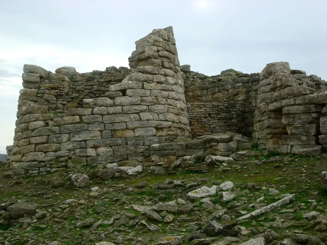 Rovine del nuraghe Su Mulinu, un'antica torre di pietra con mura ciclopiche e un paesaggio circostante roccioso e verdeggiante.