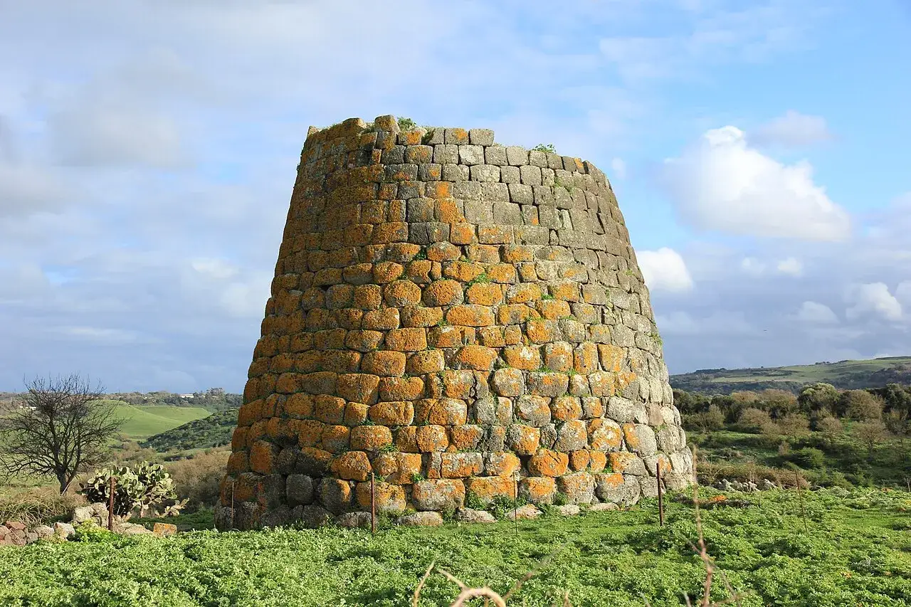 Antico nuraghe sardo, testimone silenzioso di antiche guerre. La sua mole imponente si staglia contro un cielo azzurro e nuvoloso, circondato da un paesaggio verdeggiante.