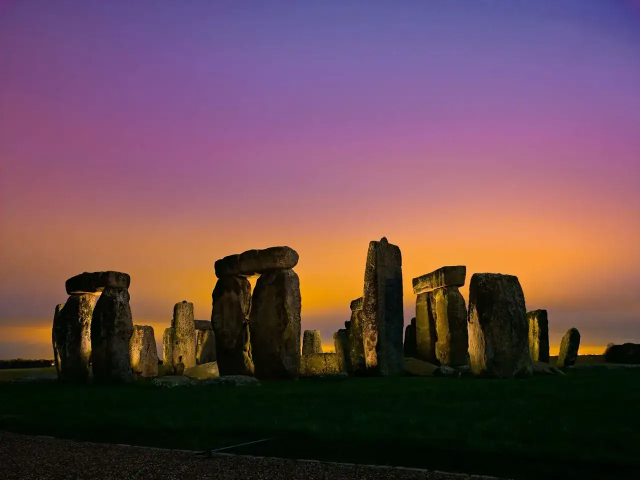 Antico cromlech illuminato da un tramonto viola e arancione, le sue pietre millenarie emergono dall'erba.