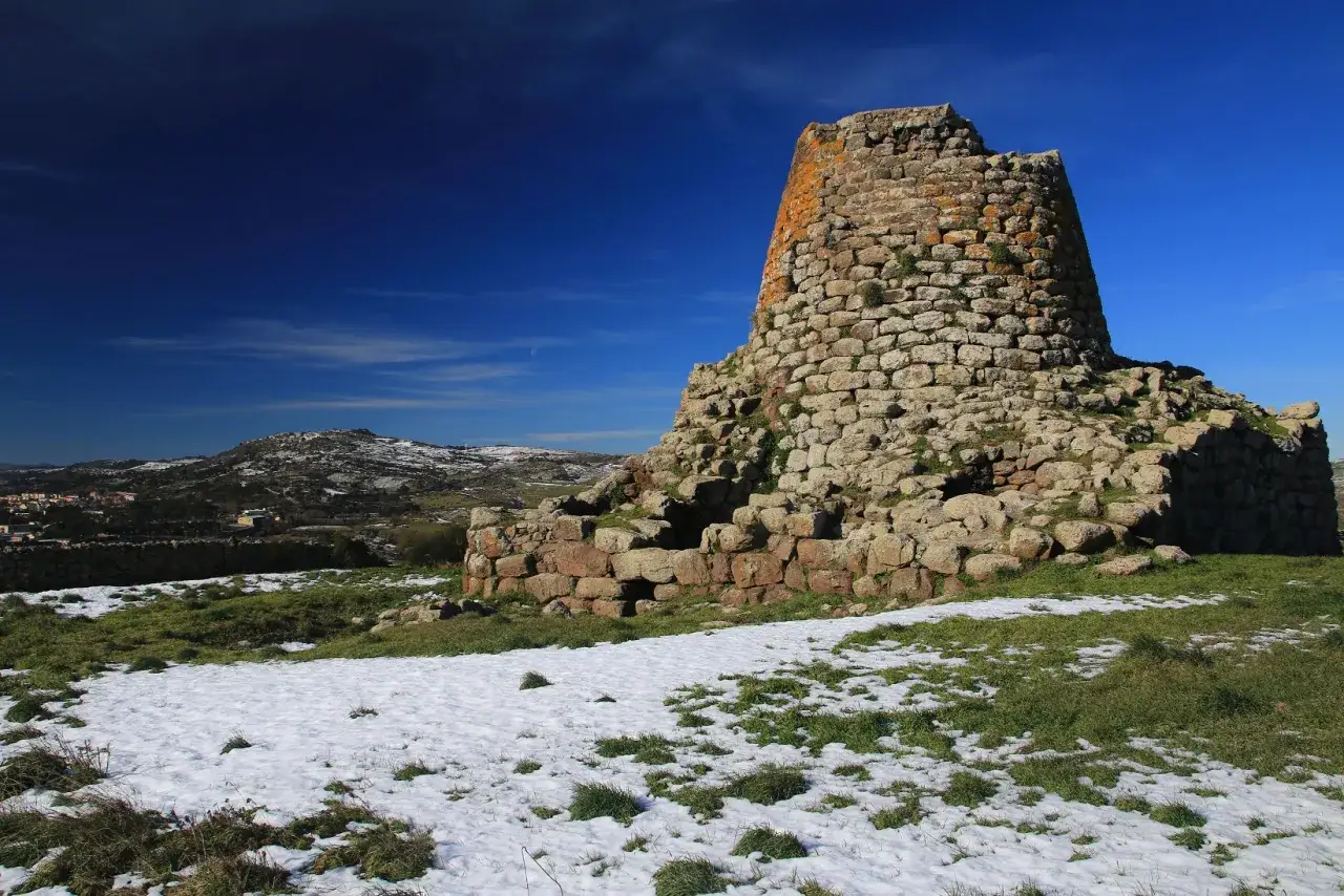 Il nuraghe Santa Barbara a Villanova Truschedu, imponente struttura in pietra, si erge su un paesaggio innevato sotto un cielo blu intenso.