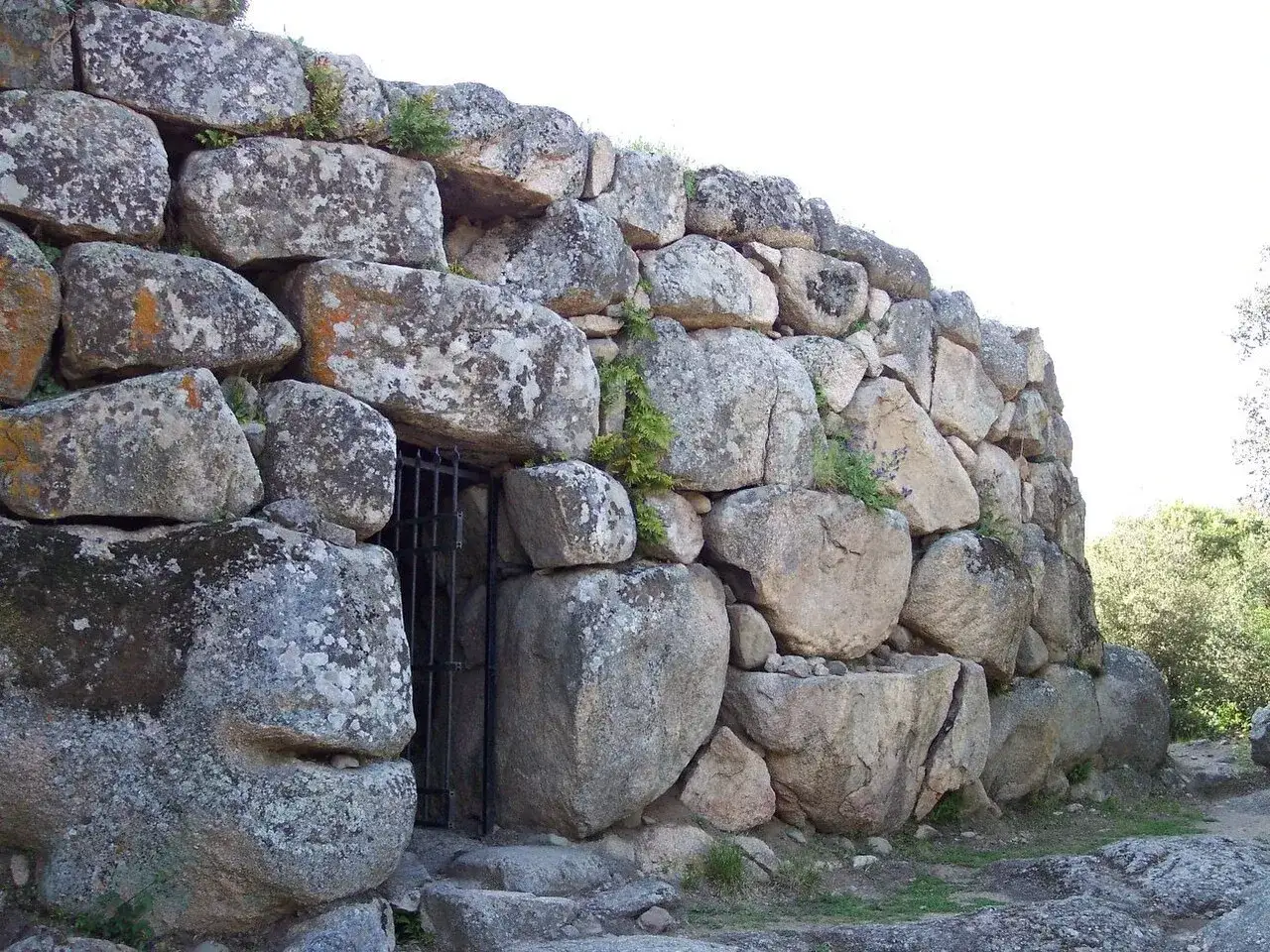 Ingresso di un nuraghe, con massi ciclopici e una porta in ferro. Un volto scolpito nella pietra sembra sorvegliare l'antico nuraghe.