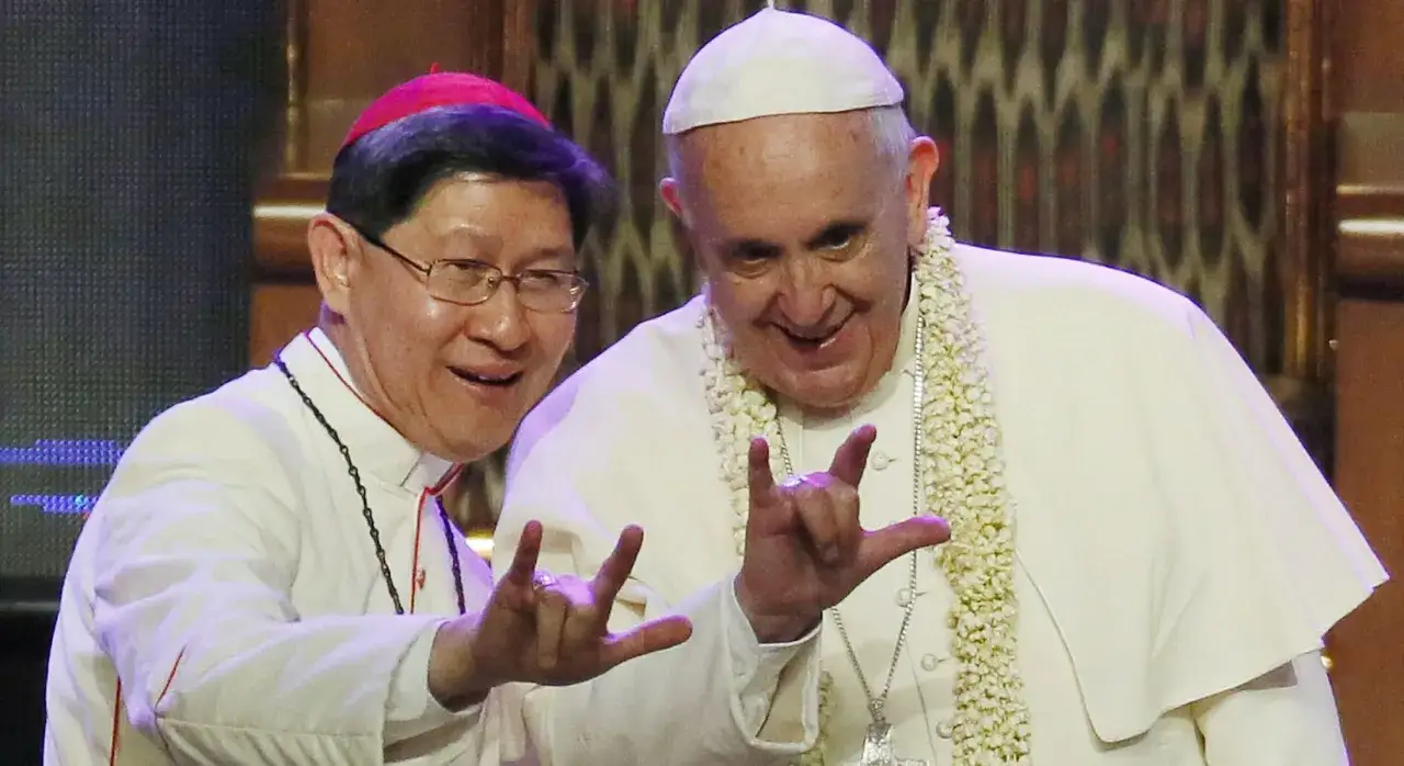 Papa Francesco e un vescovo sorridono facendo il segno delle corna a tre dita, un gesto che per alcuni ha un significato di benedizione o augurio.