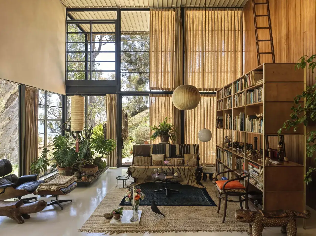 Interno della Eames House con grandi finestre, piante rigogliose, mobili iconici e una libreria piena di libri e oggetti.
