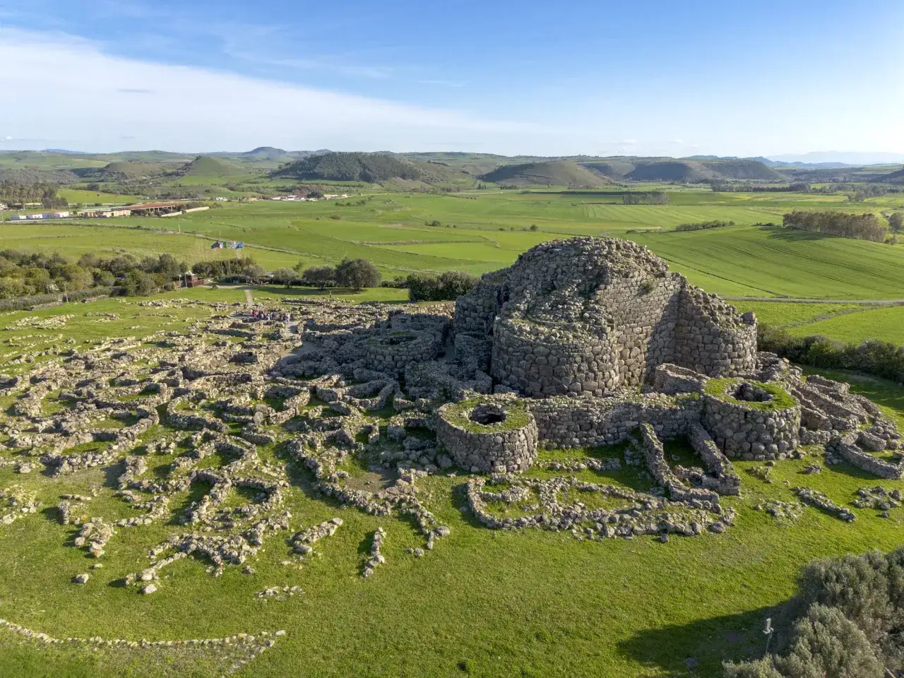 Antichi nuraghi in Sardegna, imponenti torri di pietra circondate da un villaggio di rovine, su un paesaggio verdeggiante sotto un cielo azzurro.