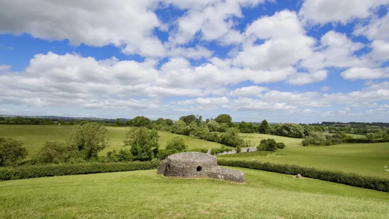 Antica struttura circolare in pietra, forse un sito archeologico di Bru na Boinne, immersa in un paesaggio verdeggiante sotto un cielo azzurro con nuvole bianche.
