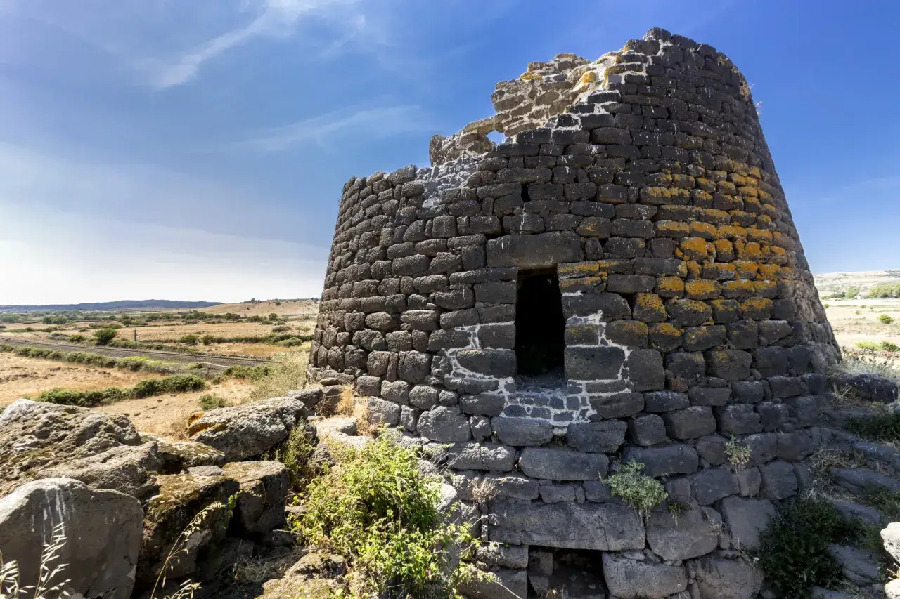 Un antico nuraghe in pietra scura, parzialmente crollato, si erge su un paesaggio arido sotto un cielo azzurro.