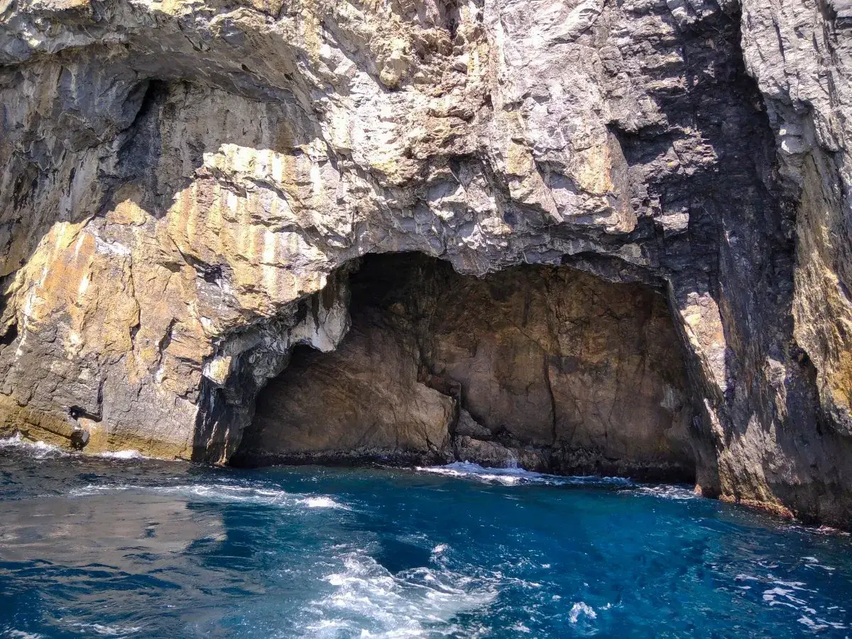 Ingresso della grotta dei colombi, con rocce scure e dorate che si tuffano nel mare blu intenso.