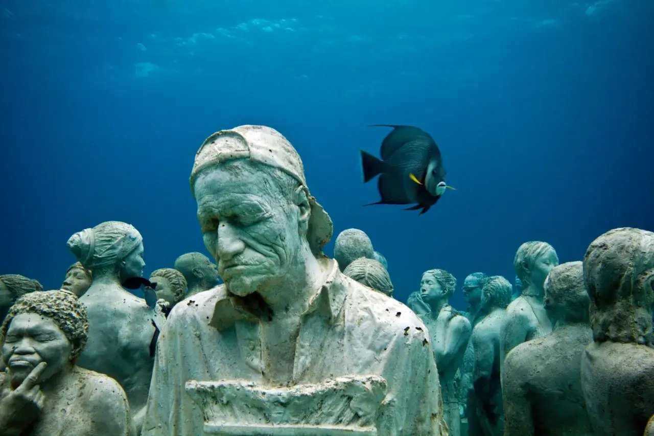 Statue giganti mare, un pesce nuota tra figure di pietra che emergono dal blu profondo.