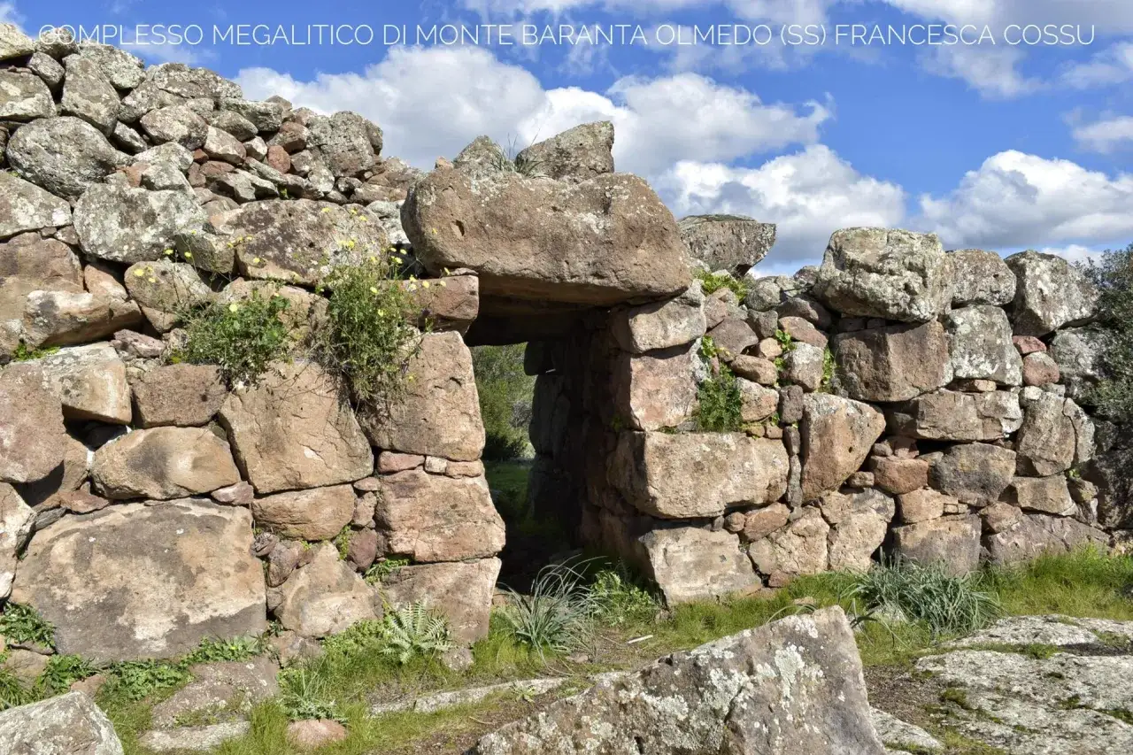 Antico ingresso del complesso megalitico di Monte Baranta, con massi imponenti e vegetazione rigogliosa sotto un cielo azzurro.