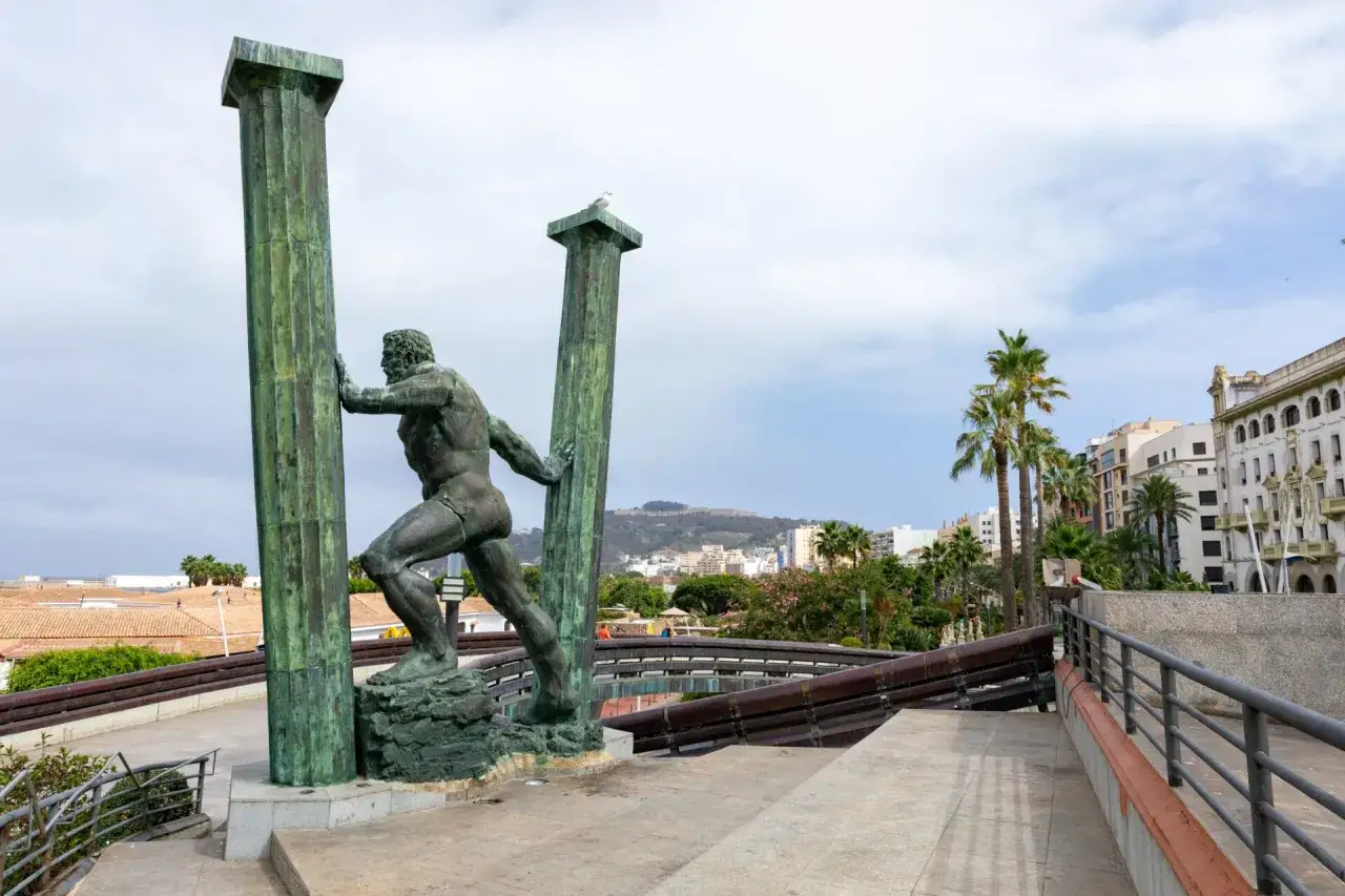 Statua di Ercole che sorregge le colonne, simbolo dei limiti del mondo antico. Sullo sfondo, palme e edifici.