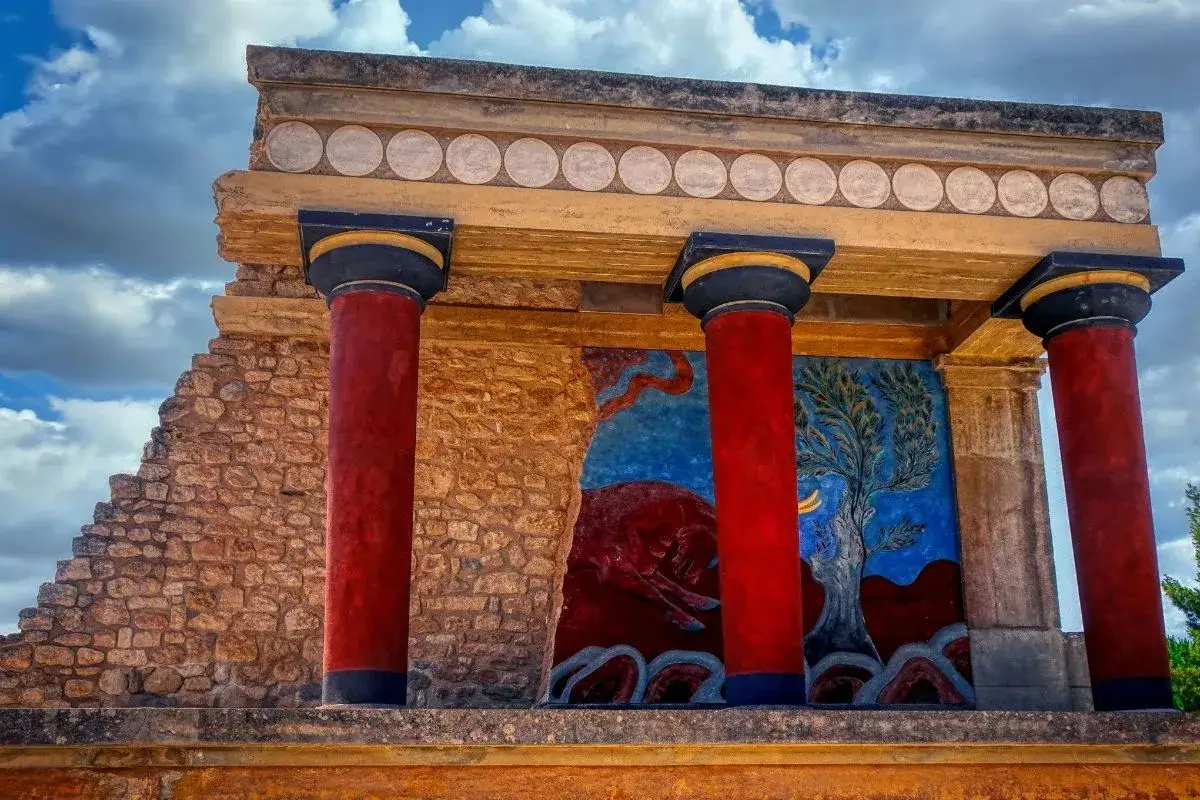 Resti di un palazzo minoico con colonne rosse e affreschi che raffigurano scene di vita, forse un toro, e un albero, evocando l'arte dei minoici e micenei.