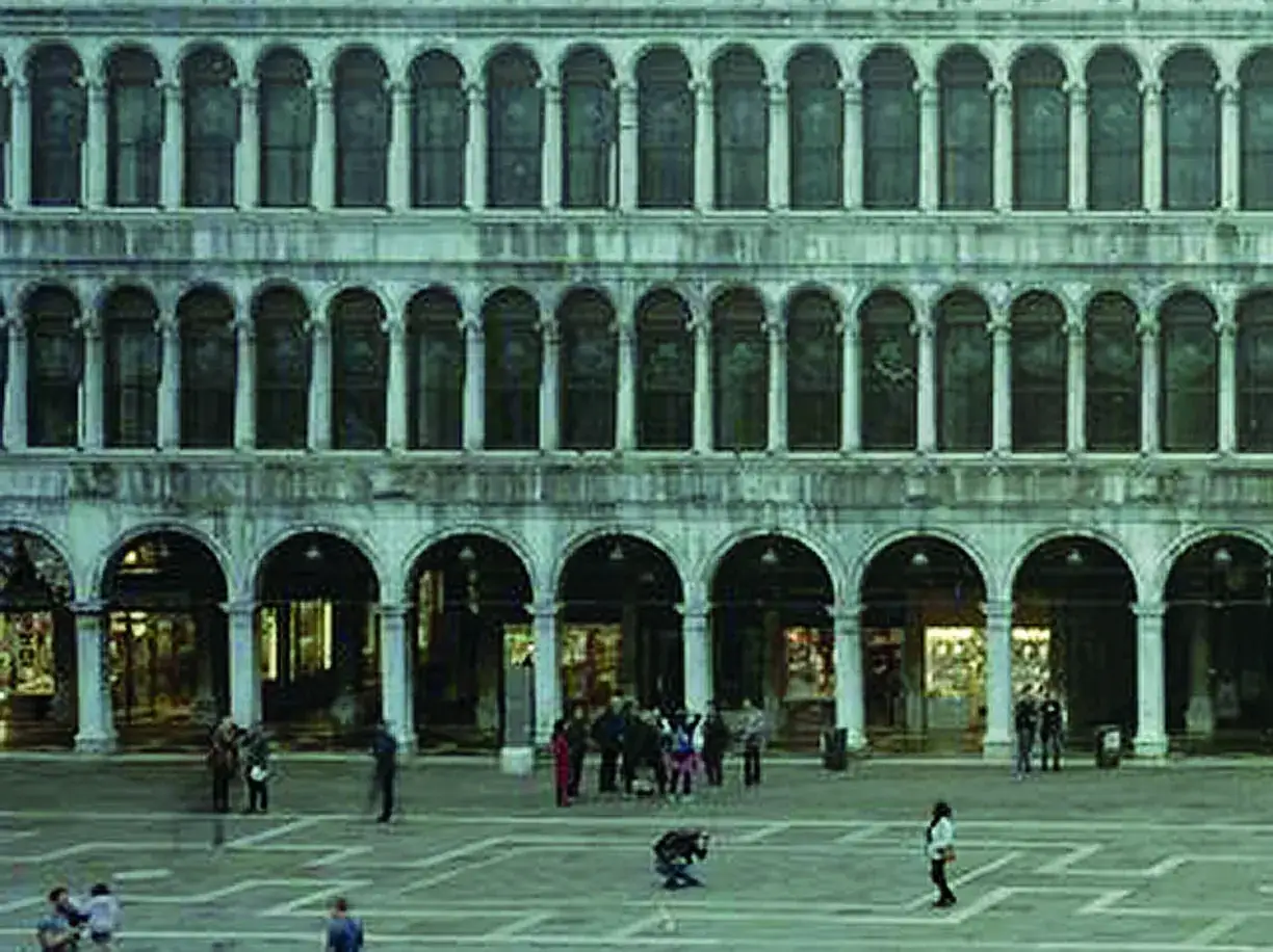 Un imponente palazzo con arcate si affaccia su Piazza San Marco, dove alcuni turisti passeggiano.