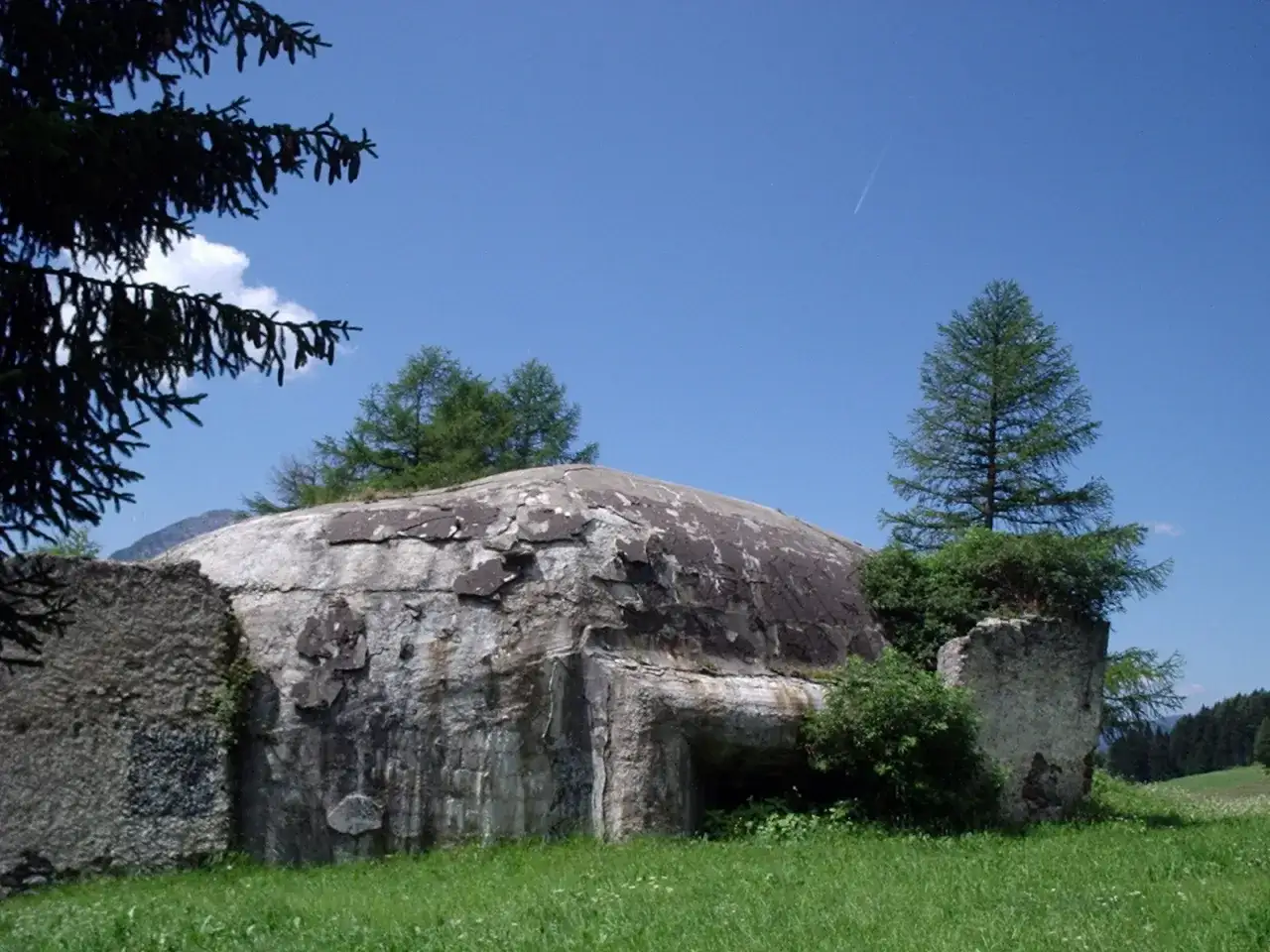 Un bunker del vallo alpino del littorio, con vegetazione che cresce intorno, sotto un cielo azzurro con una scia di aereo.
