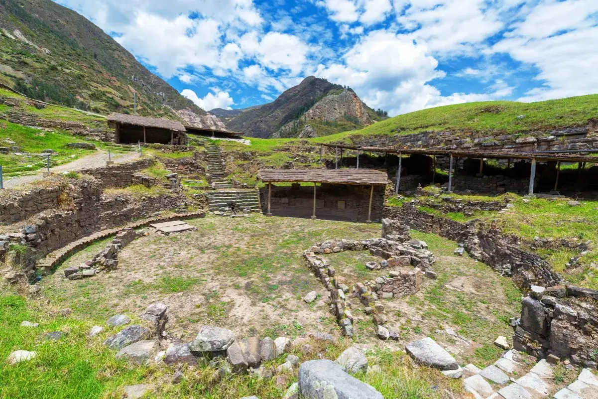 Rovine di Chavin de Huantar, con antiche strutture in pietra e tetti di paglia, incorniciate da verdi colline e un cielo azzurro con nuvole bianche.