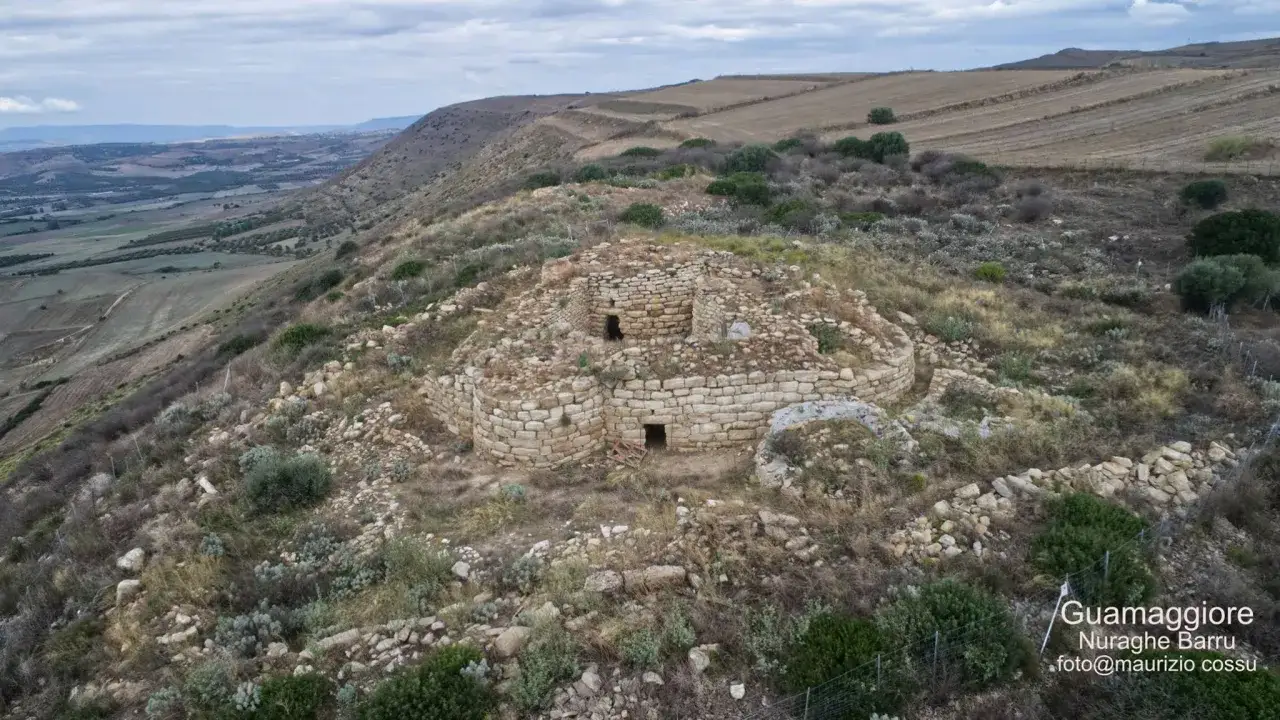 Antica struttura in pietra del nuraghe Barru, immersa in un paesaggio collinare sardo con campi coltivati all'orizzonte.