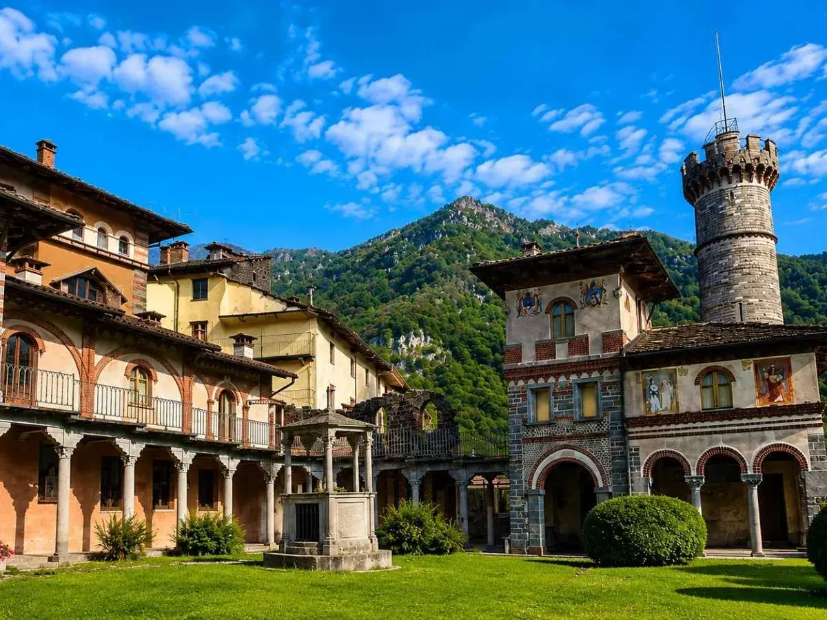 Cortile con pozzo e torre, circondato da edifici storici con affreschi, immerso in un paesaggio montano. Un luogo che evoca un'atmosfera di rosazza esoterica.